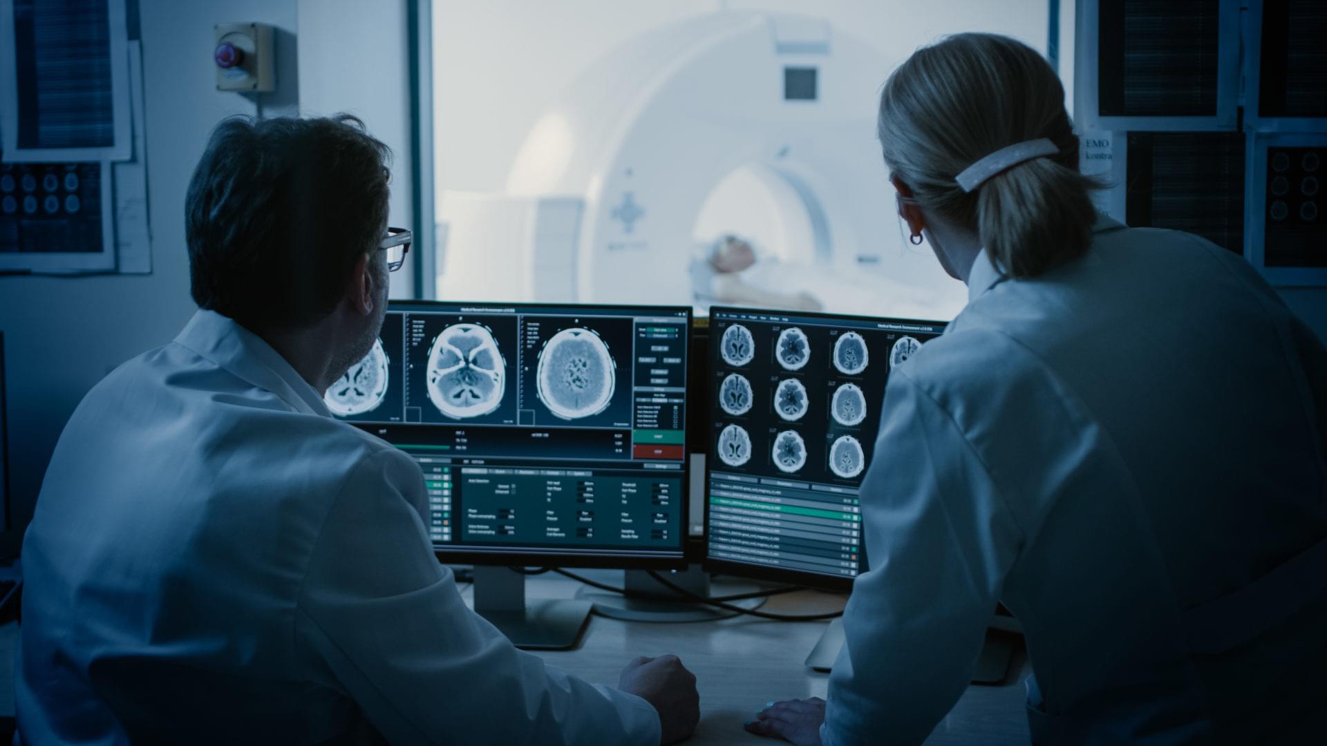 Im Kontrollraum besprechen Arzt und Radiologe die Diagnose, während sie die Prozedur beobachten und die Ergebnisse der Hirnscans überwachen. Im Hintergrund unterzieht sich der Patient einem MRT- oder CT-Scan-Verfahren.