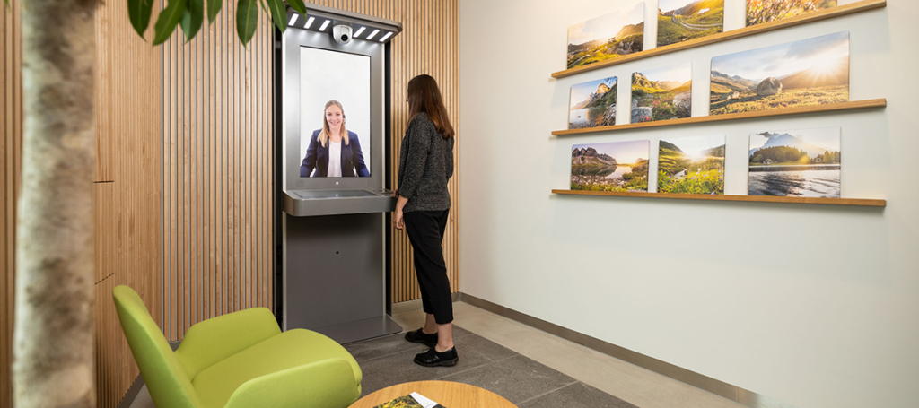 Frau steht vor einem Videoserviceterminal. Auf dem Bildschirm ist eine Beraterin zu sehen.